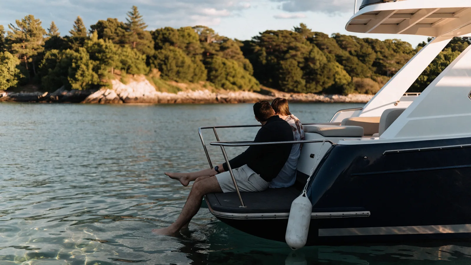 Ein Paar sitzt am Abend auf der Badeplattform seiner Motoryacht in einer stillen Bucht, die Füße im Wasser