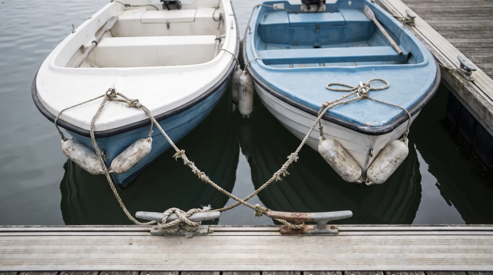 Two small open motorboats lying side by side at a timber pontoon, their bow lines crossed and made fast on two cleats.