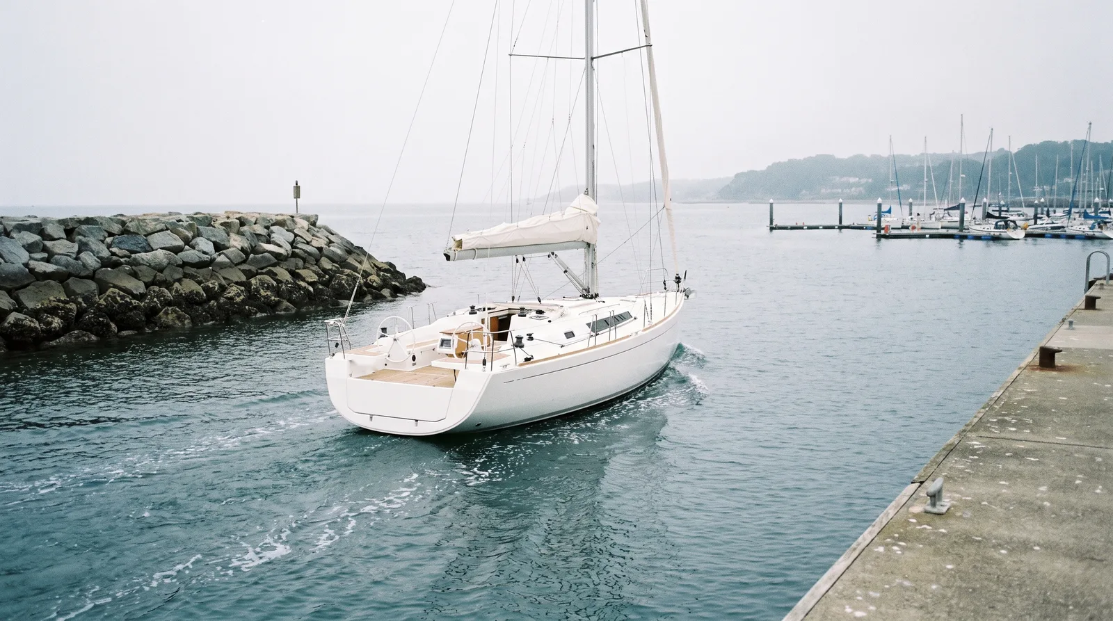 Eine weiße Segelyacht mit eingerollten Segeln läuft unter Motor an einer Steinmole vorbei aus dem Hafen, im Hintergrund Schwimmstege mit Booten und diesige Hügel.