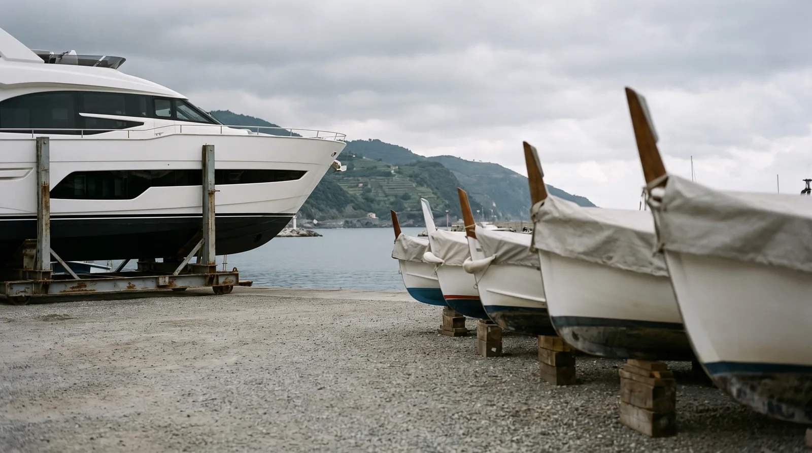 A boatyard quay on the Ligurian coast: on the left a new white motor yacht on a rusty steel transport cradle, on the right a row of small wooden boats on blocks under grey covers, with calm harbour water and terraced hills under an overcast sky.
