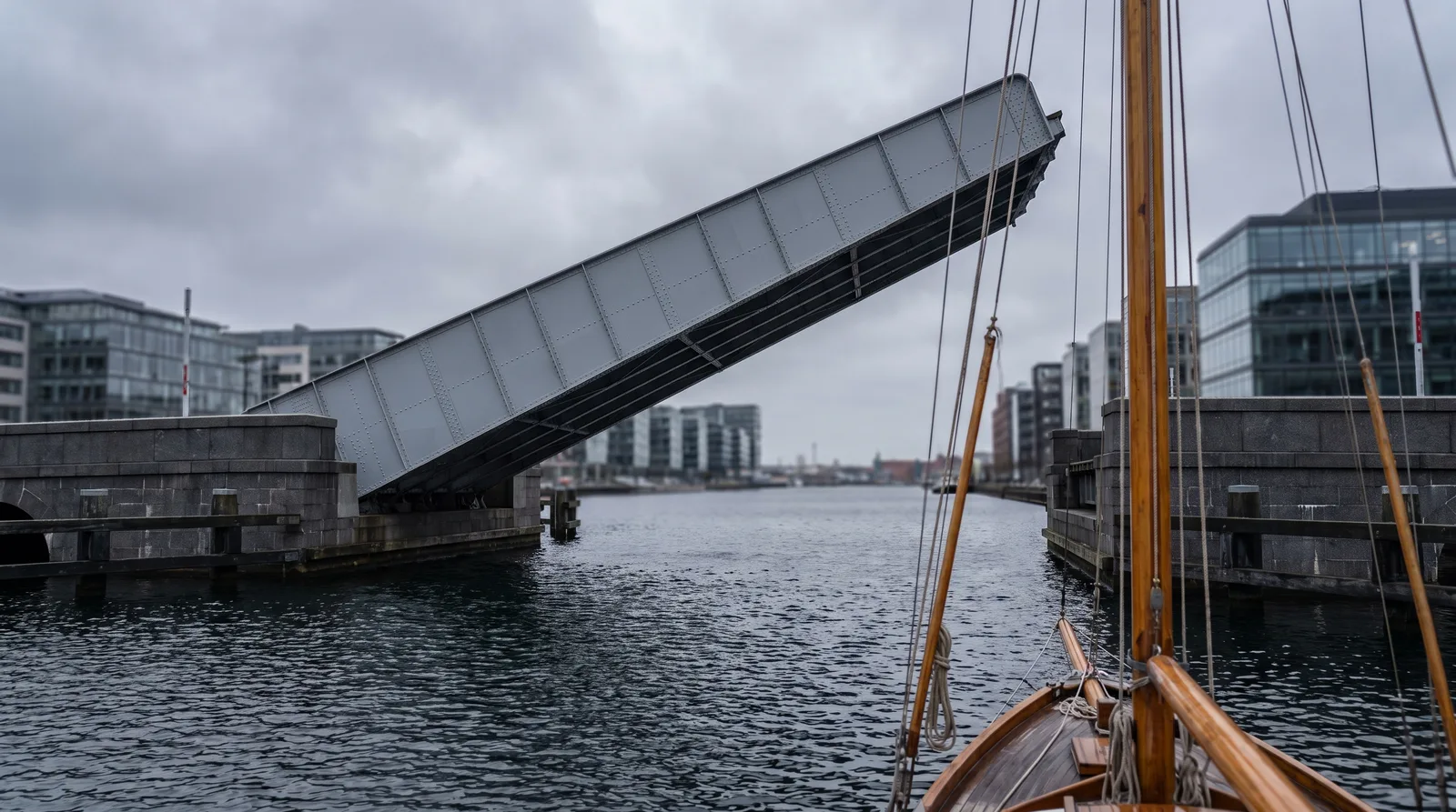 Die hochgestellte Klappe einer stählernen Hafenbrücke über dunklem Wasser, im Vordergrund unscharf der Holzmast eines kleinen Segelbootes.