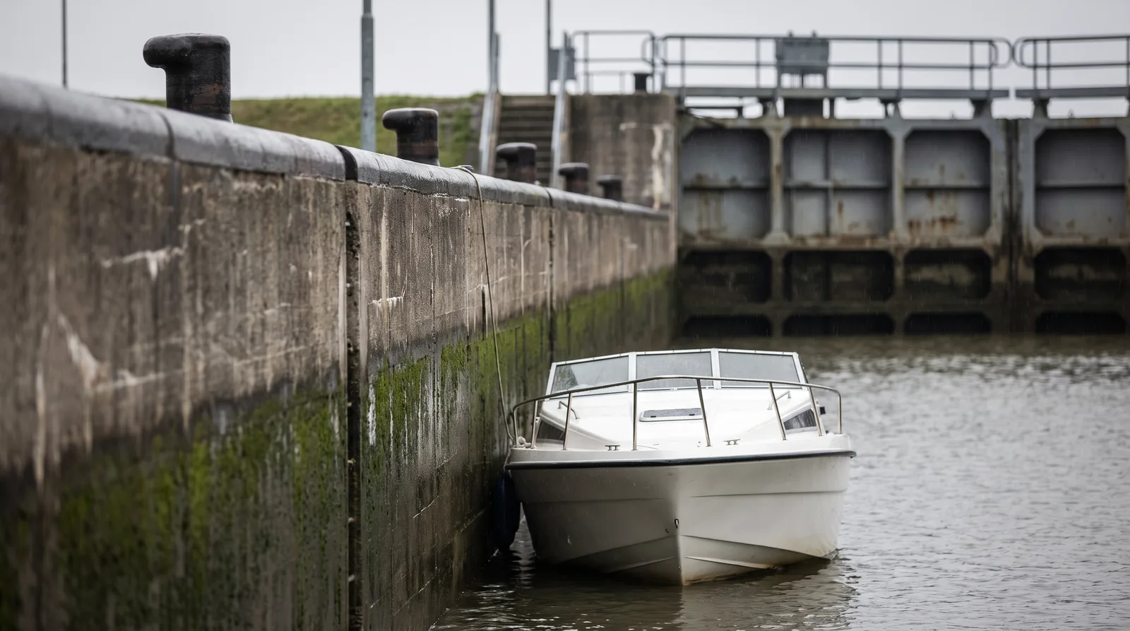 A small white motor cruiser lies against the mossy concrete wall of a lock chamber under grey light, with heavy iron bollards on the coping and the lock gate behind.