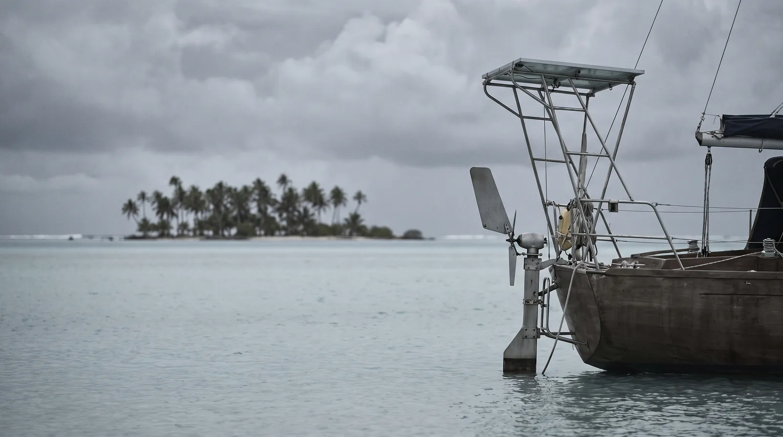 The stern of a cruising yacht lying at anchor in a wide lagoon, with a windvane self steering gear on the transom and a solar panel on a stainless arch. A low island of coconut palms sits under a heavy overcast sky.