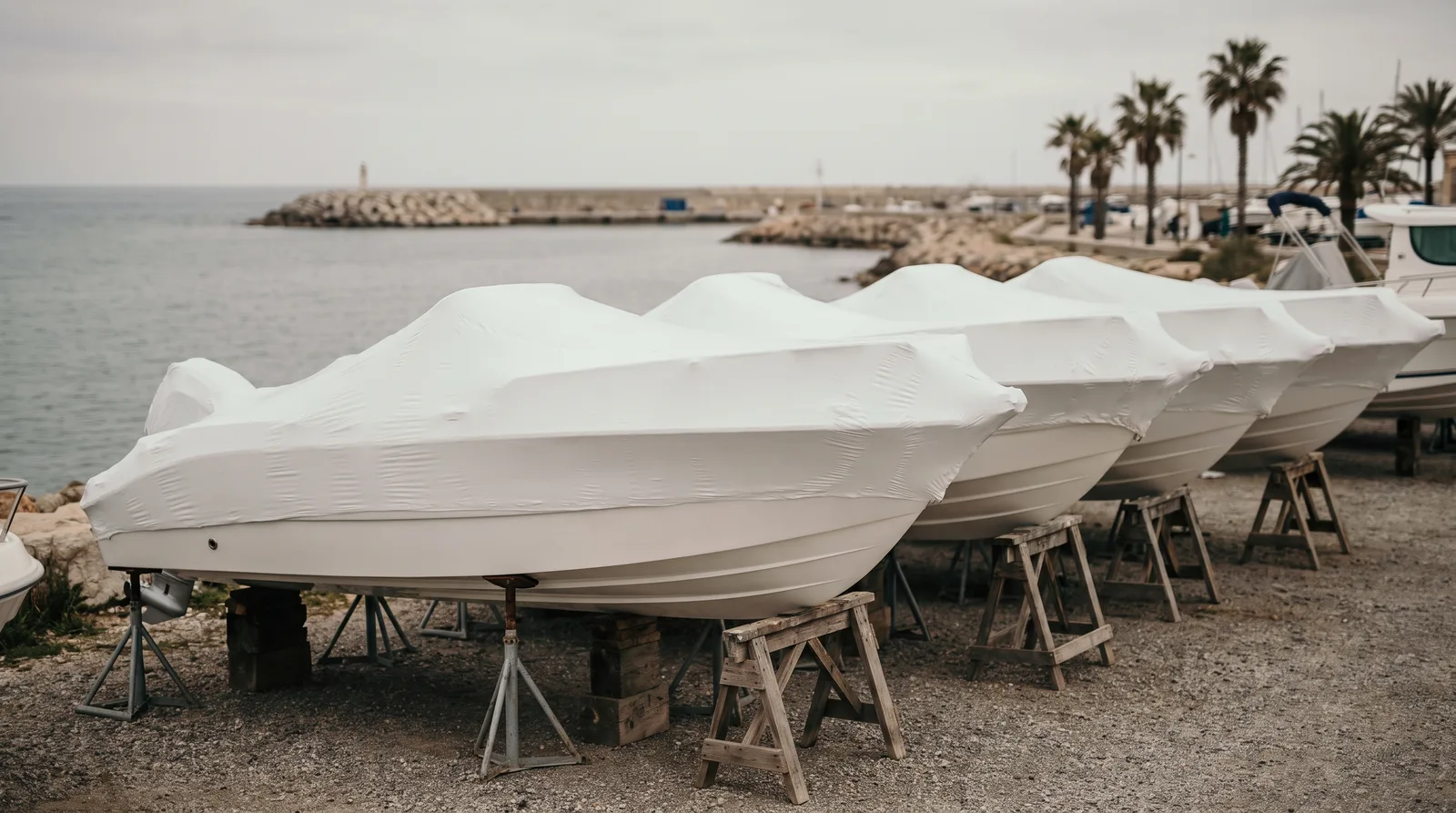 Eine Reihe neuer Motorboote in weißer Schrumpffolie auf Holzböcken und Stützen auf einem Kiesplatz am Meer, dahinter eine Mole und Palmen unter grauem Himmel.