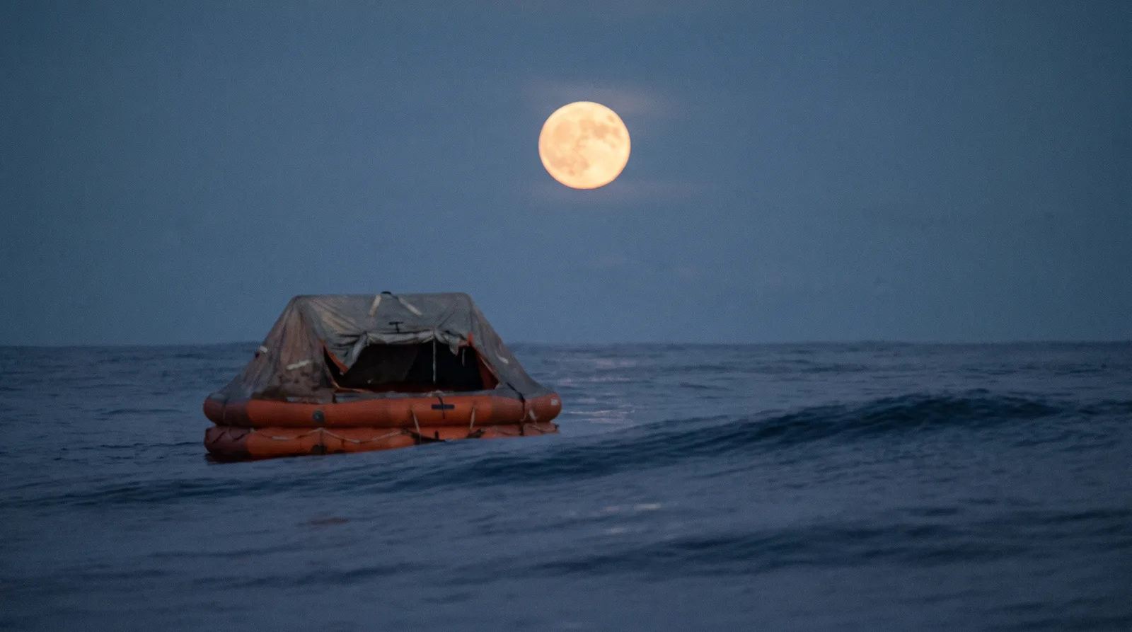 An orange canopied life raft drifting alone on a long swell under a full moon