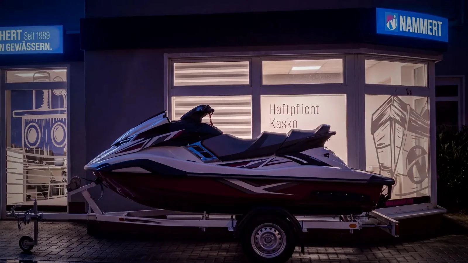 Das Büro in der Karl-Marx-Straße 4 in Königs Wusterhausen am Abend, die beleuchteten Schilder über den Schaufenstern, darin das gezeichnete Ruderboot und der Schlepper, davor ein Wasserfahrzeug auf einem Trailer