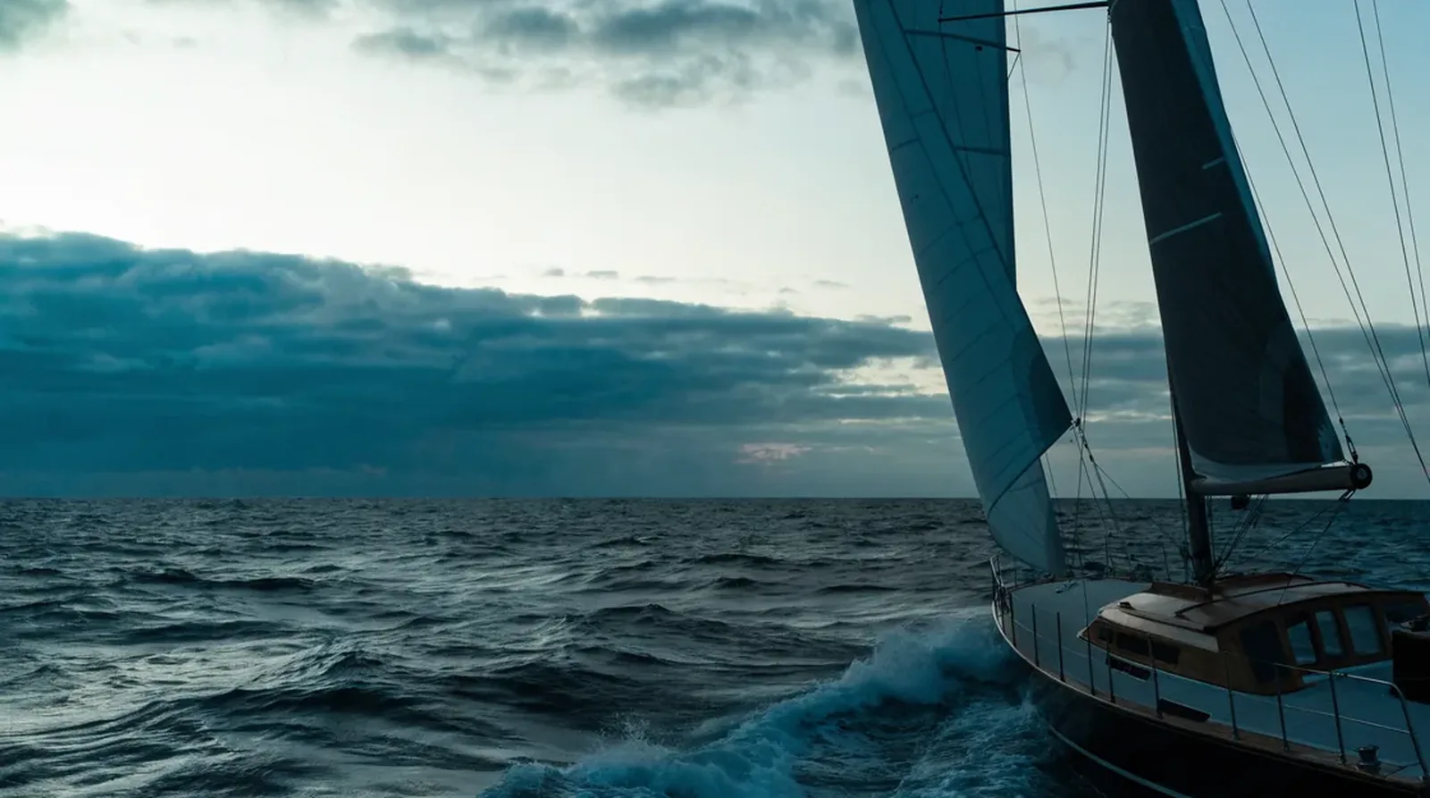 Segelyacht unter Segeln auf offener, bewegter See am Abend, Großsegel und Vorsegel stehen vor einem wolkigen Himmel, am Bug bricht Gischt.