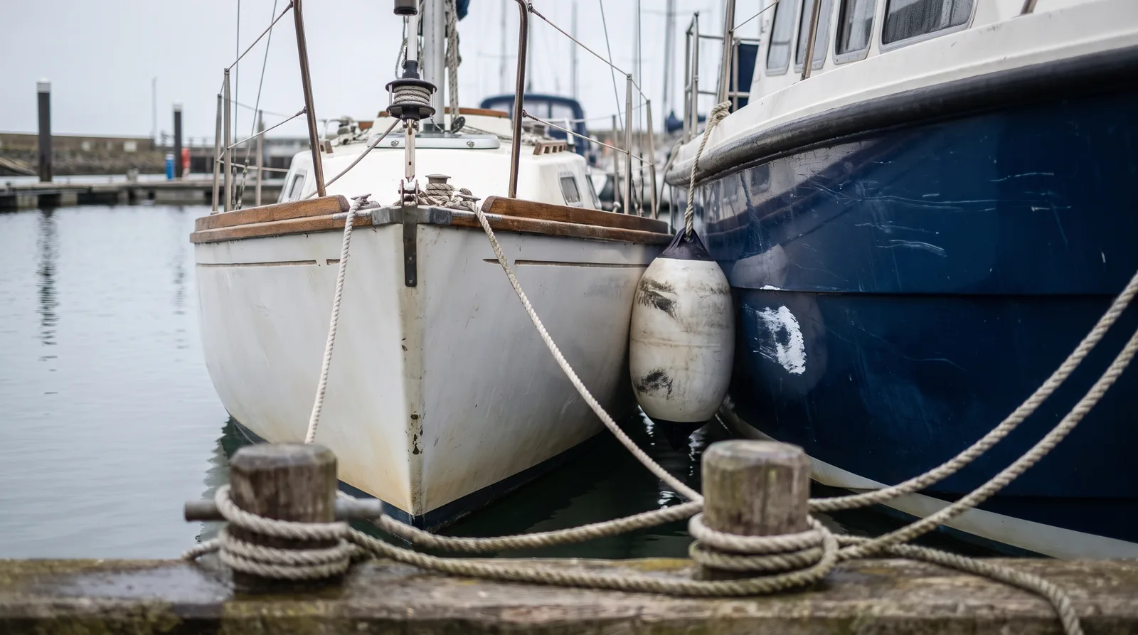 Der weiße Bug einer Segelyacht dicht an der dunkelblauen Bordwand eines Motorboots, dazwischen ein verschrammter Fender, an der Bordwand eine frische Schramme, vorn Poller mit Festmacherleinen am Holzsteg, im grauen Tageslicht