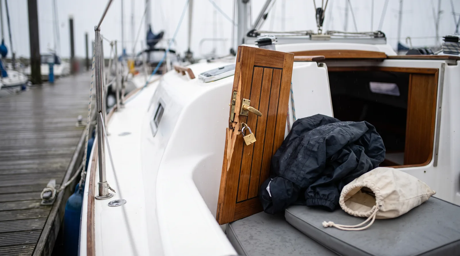 Am Niedergang eines gepflegten weißen Segelboots in einem Hafen steht das hölzerne Steckschott schief, am Beschlag hängt ein Vorhängeschloss, das Holz daneben ist frisch gesplittert, auf der Cockpitbank liegen eine dunkle Regenjacke und ein leerer Stoffbeutel