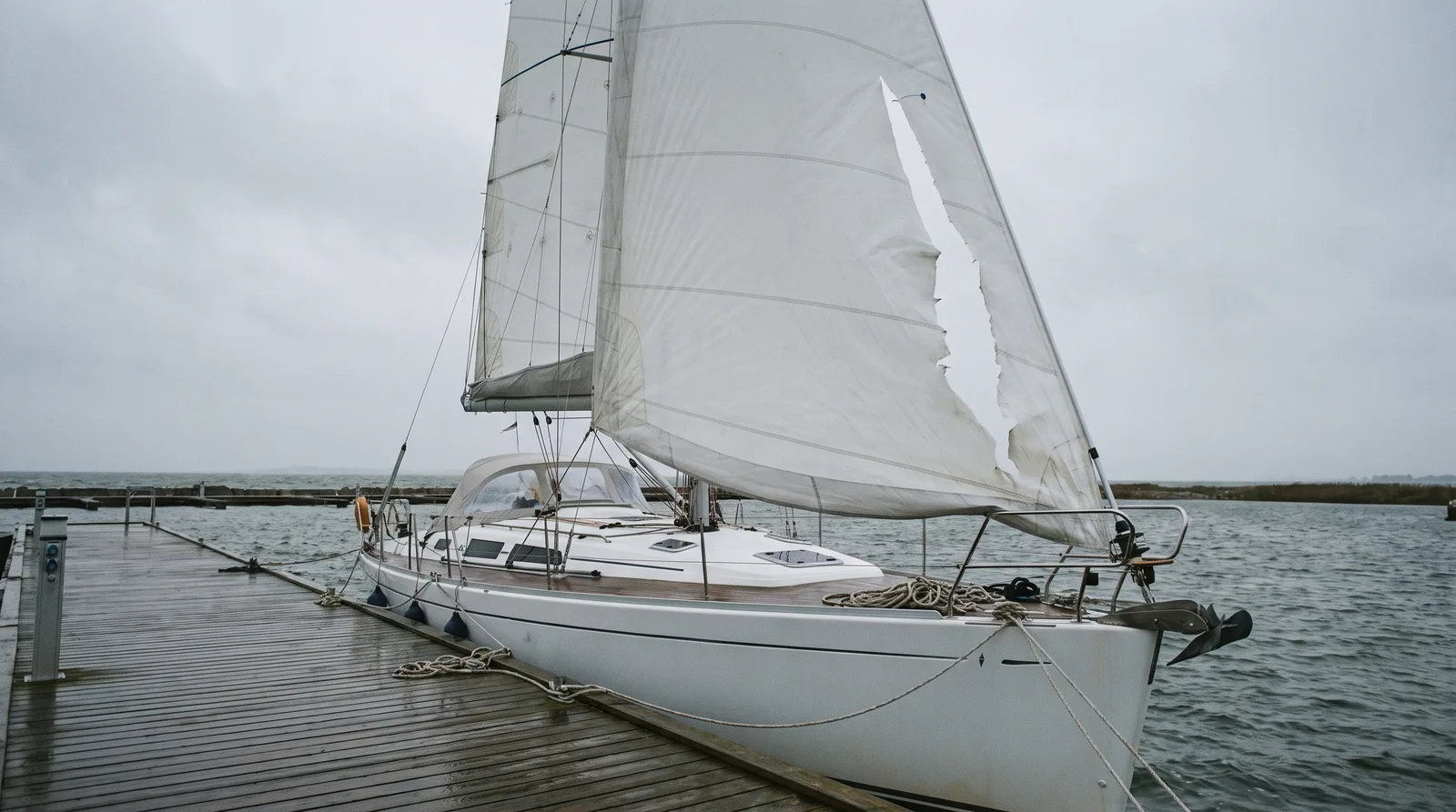 Eine weiße Segelyacht liegt an einem nassen Holzsteg an der Ostsee, die halb abgerollte Genua hat einen langen Riss quer durch mehrere Tuchbahnen, grauer Himmel und aufgewühltes Wasser