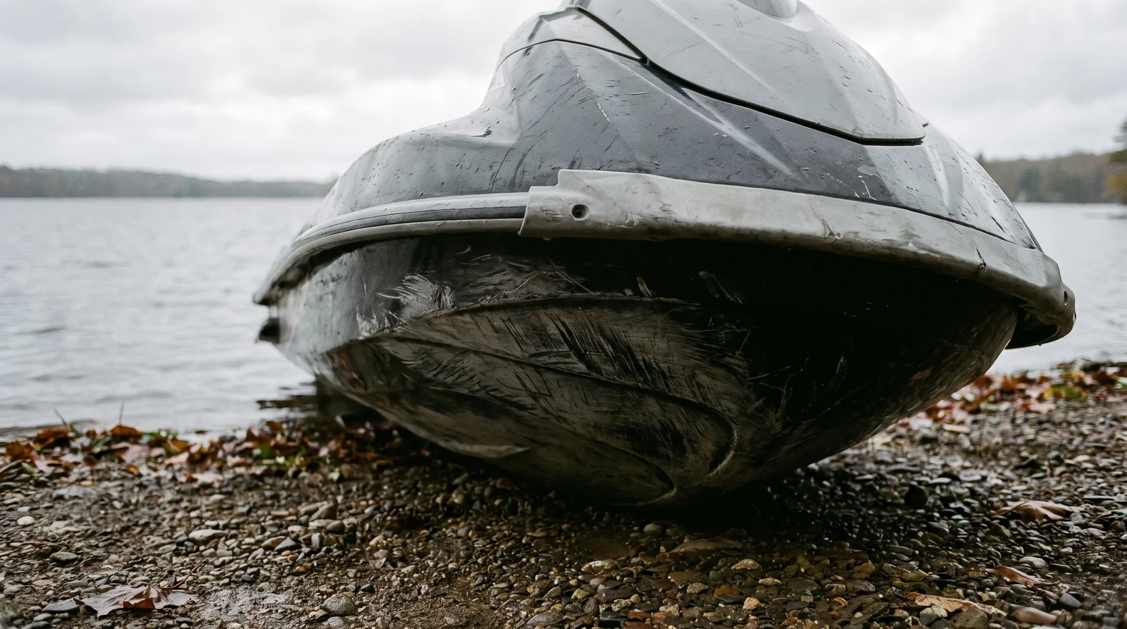 Bow of a jet ski with scratches on the hull, resting on a gravel shore under an overcast sky.