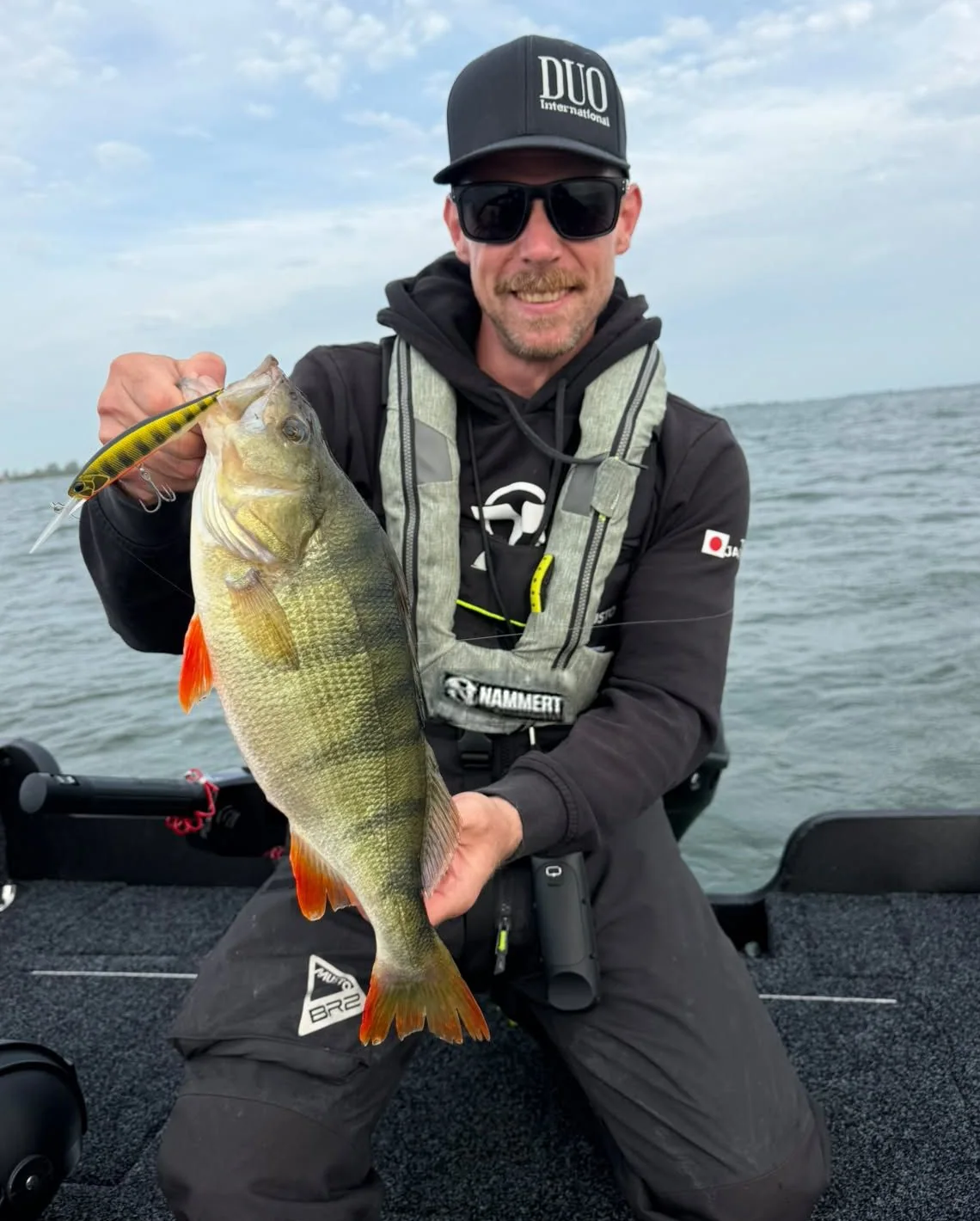 Angler kneeling in a boat holding a large perch, the lure beside it, the life vest reading NAMMERT