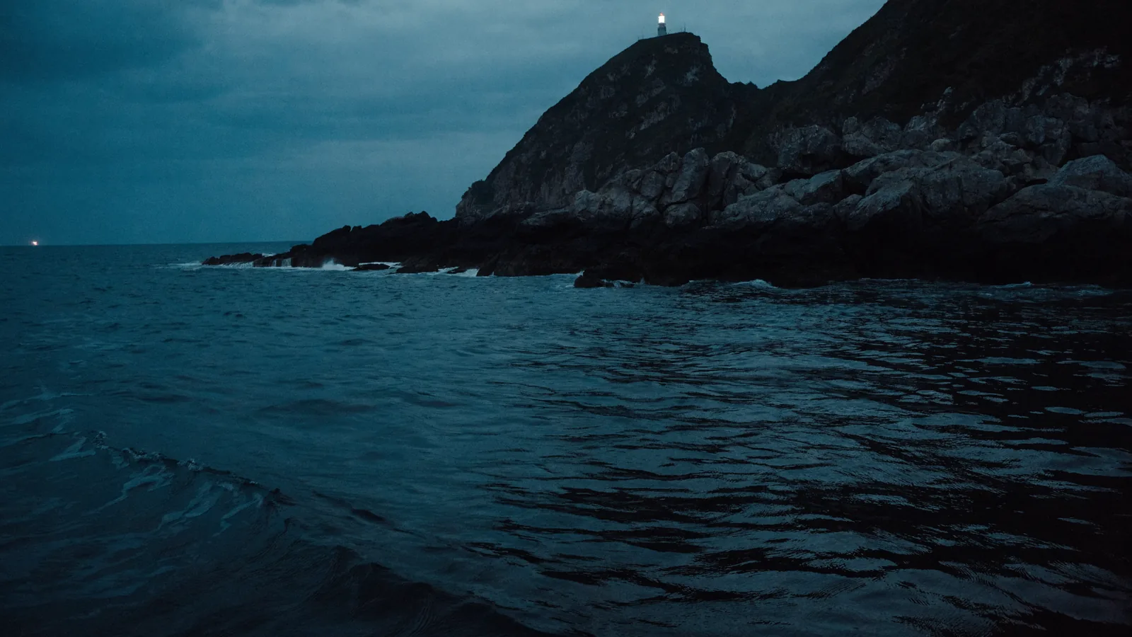 Felsküste mit Leuchtturm vom Wasser aus in der Dämmerung