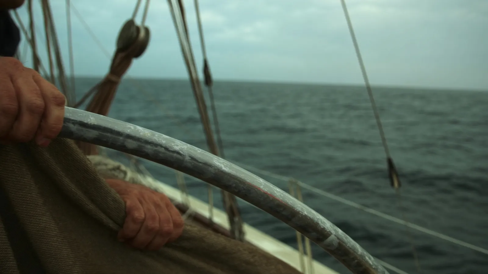 A hand on the wheel of a cruising yacht, open water ahead under an overcast sky