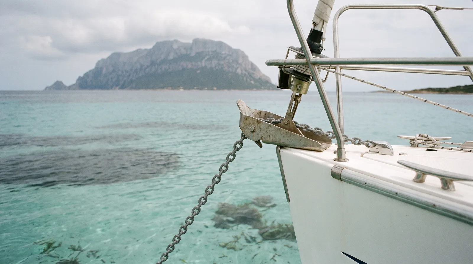 Bug einer Segelyacht mit Ankerkette in türkisem Wasser über hellem Sand, dahinter die Insel Tavolara