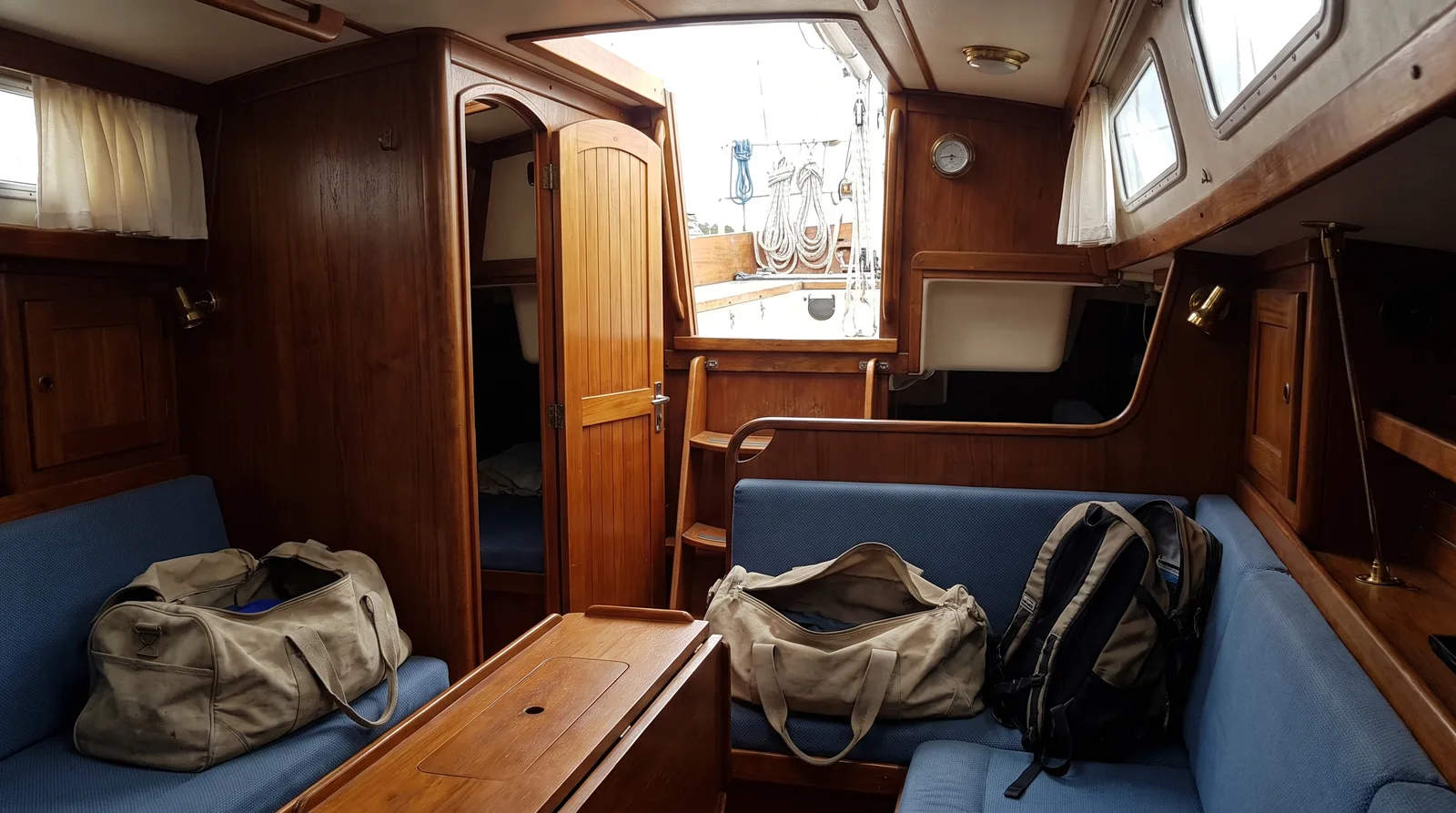 Salon einer Segelyacht mit Holzausbau, auf den Polsterbänken offene Reisetaschen und ein Rucksack, der Niedergang steht offen