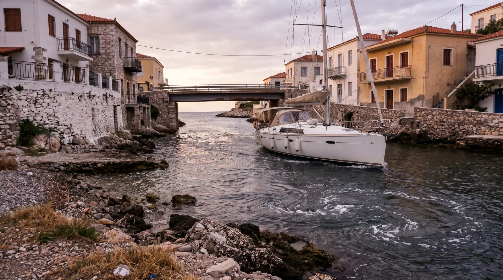Segelyacht unter Motor in einer schmalen Durchfahrt zwischen griechischen Häusern mit Balkonen, dahinter eine niedrige Brücke, auf dem Wasser Strudel der Strömung