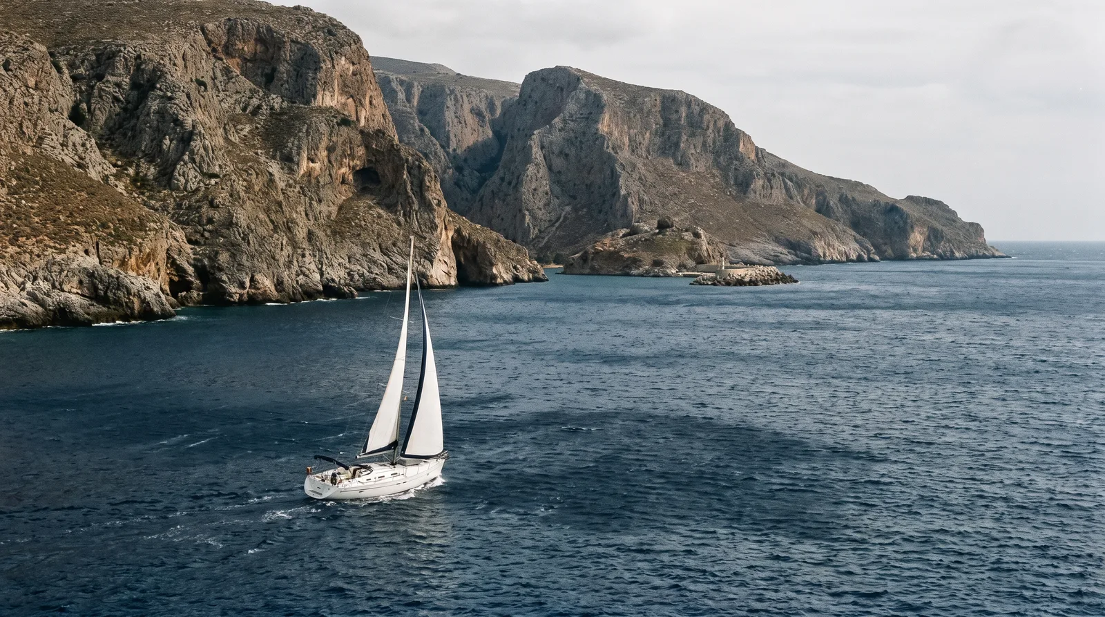 Weiße Segelyacht unter Segeln vor der steilen Felsküste mit Schluchten im Süden Kretas, dunkle Böenfelder auf dem Wasser