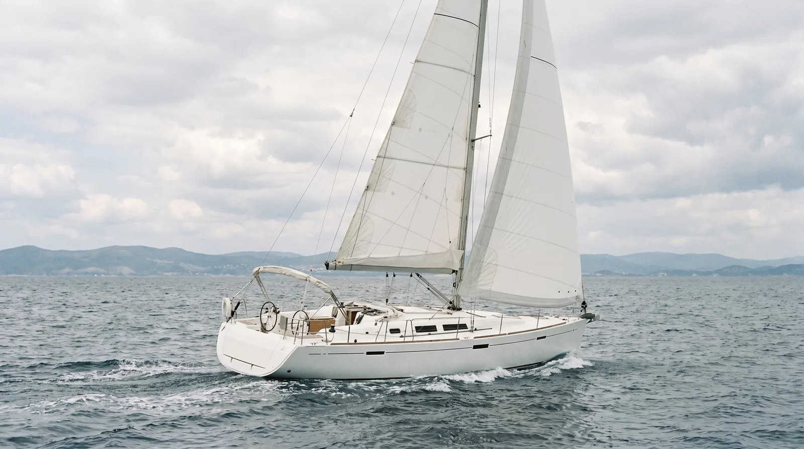 Moderne weiße Charteryacht unter vollen Segeln auf offenem Wasser mit Kielwasser und fernem Küstenstreifen