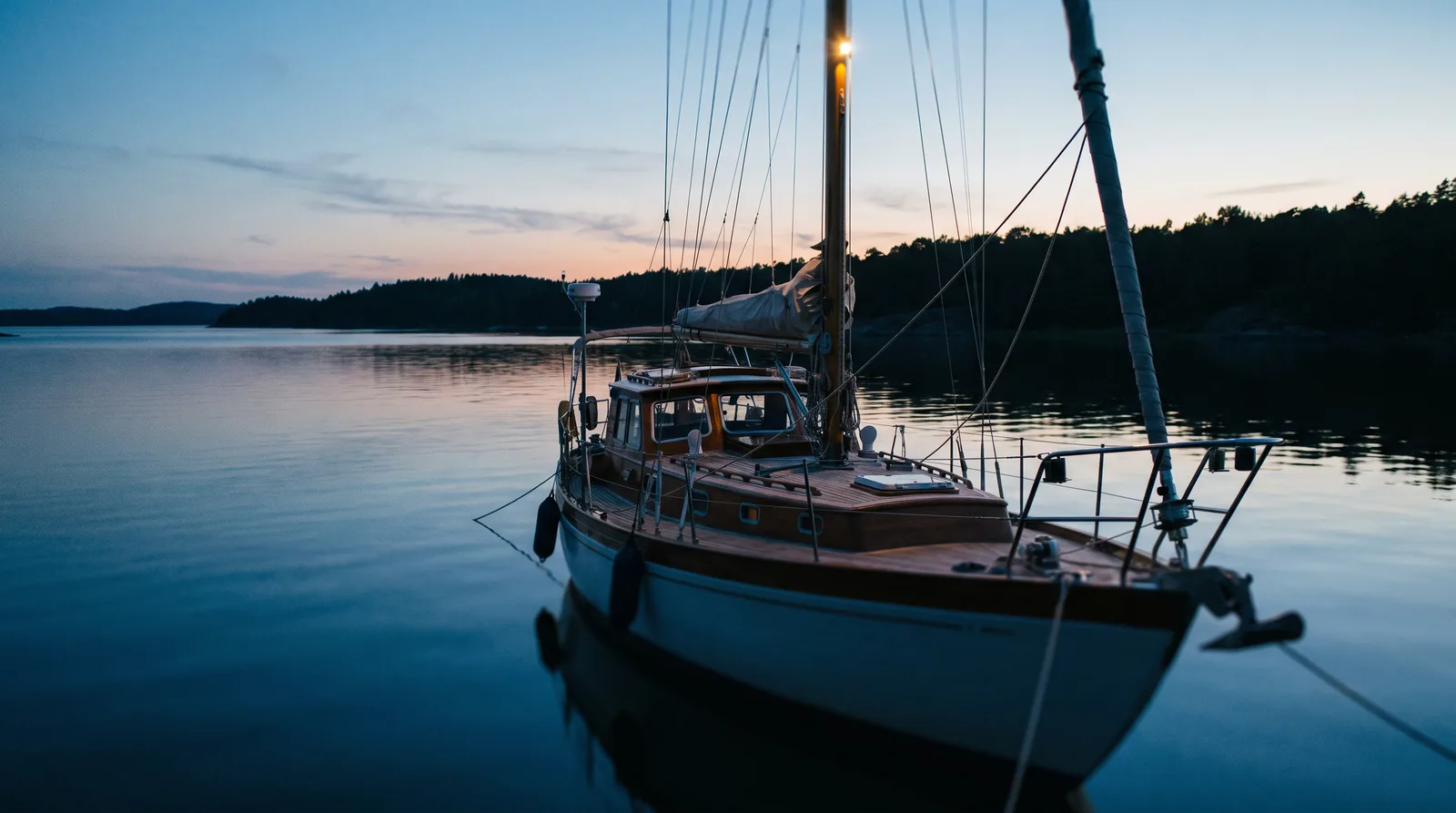 Segelyacht liegt zur blauen Stunde in einer geschützten Bucht vor Anker, Ankerlicht im Masttop, ruhiges Wasser