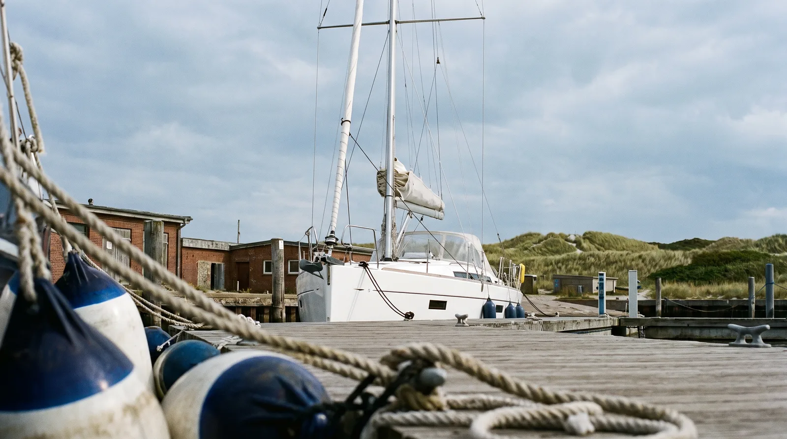 Moderne Segelyacht an einem Holzsteg in einem Ostseehafen, vorn Fender und Festmacherleinen, dahinter Backsteinhäuser und Dünen