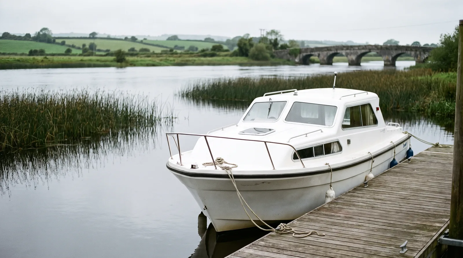 Weißer Mietkabinenkreuzer an einem Holzsteg auf einem ruhigen, breiten Fluss in Irland mit Schilf, Wiesen und Steinbrücke