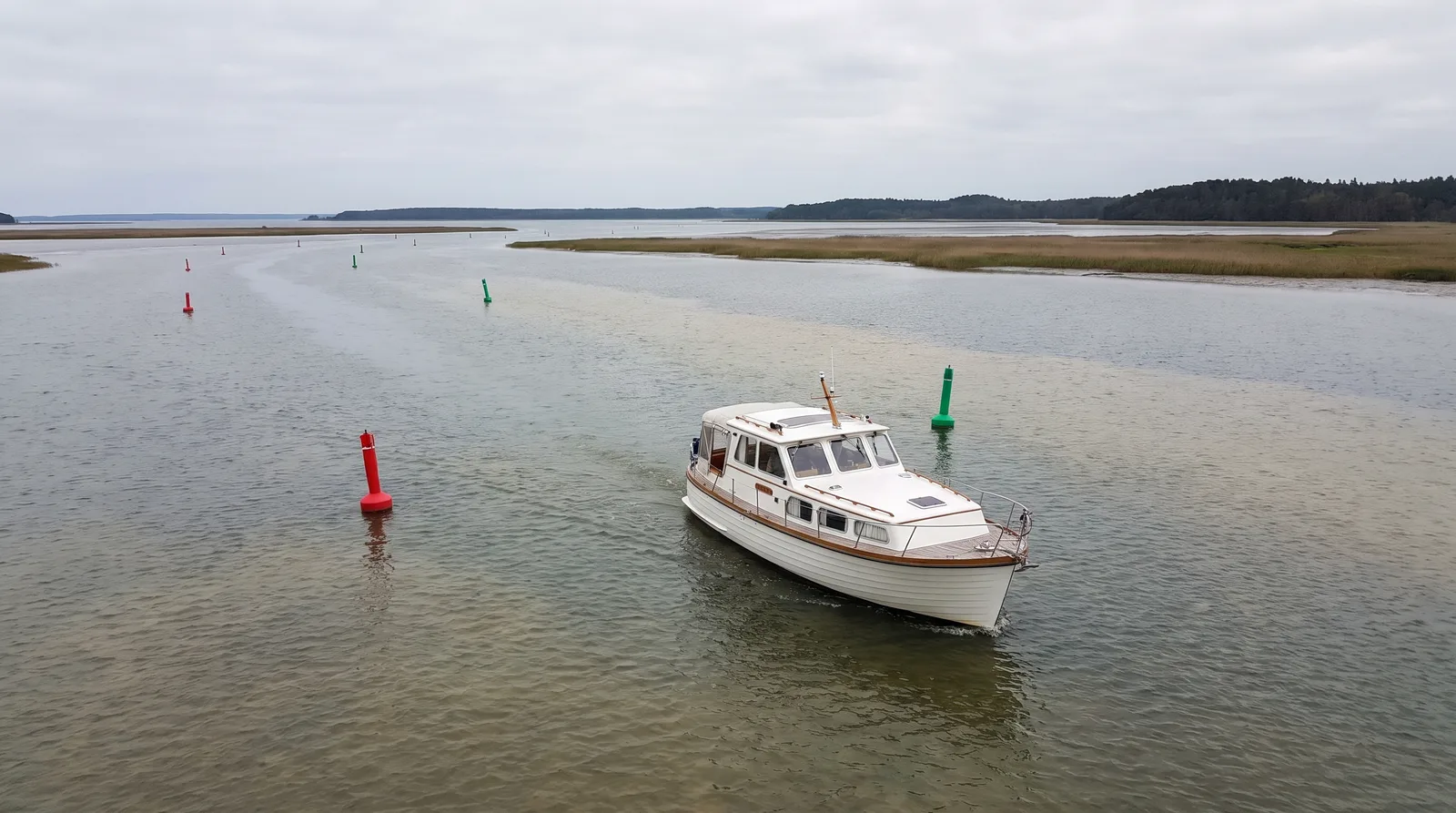 Weißes Motorboot fährt langsam zwischen roten und grünen Tonnen durch ein flaches, weites Haff mit Schilfufern und flachem Waldrand