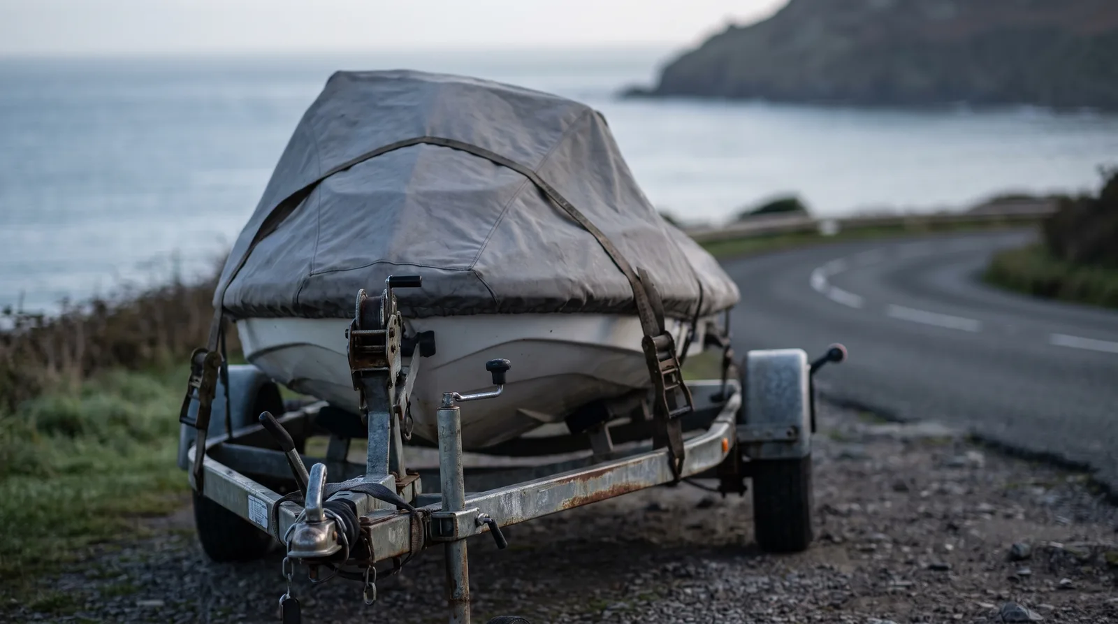 Ein abgedecktes Wassermotorrad steht festgezurrt auf einem Trailer an einer Küstenstraße, im Hintergrund das Meer