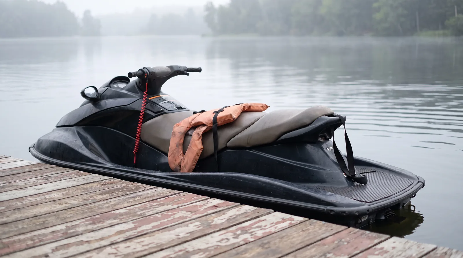Ein leerer Jetski mit Schwimmweste auf der langen Sitzbank und roter Notstoppleine am Lenker liegt an einem Holzsteg im Morgendunst