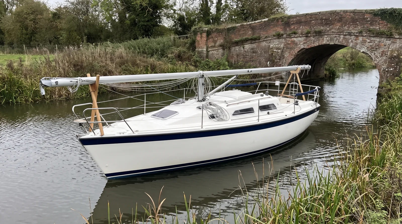 Gepflegte Segelyacht mit gelegtem Mast auf Holzböcken auf einem schmalen Fluss vor einer alten Ziegelbogenbrücke