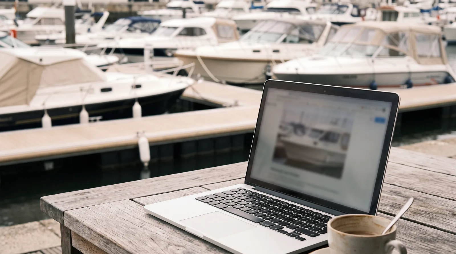 Laptop mit Bootsbörse auf einem Tisch im Hafen, im Hintergrund unscharf gepflegte Motorboote am Steg