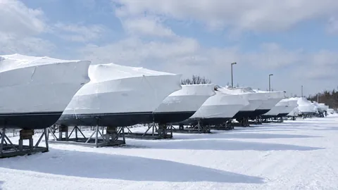 Boats on cradles under covers in a yard with snow on the ground