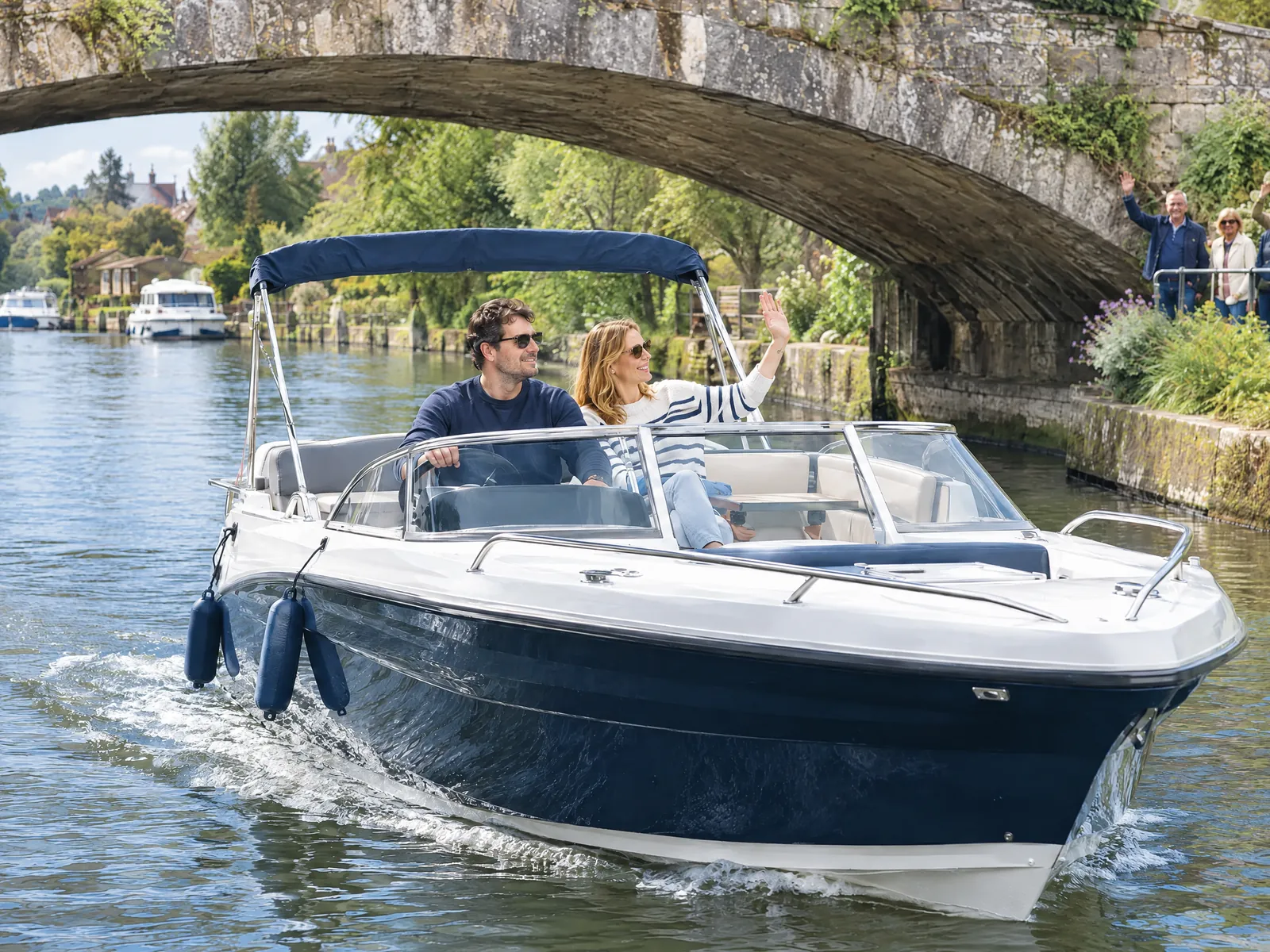 A couple cruise under an old stone bridge in a navy and white motorboat, she waves to people on the riverbank