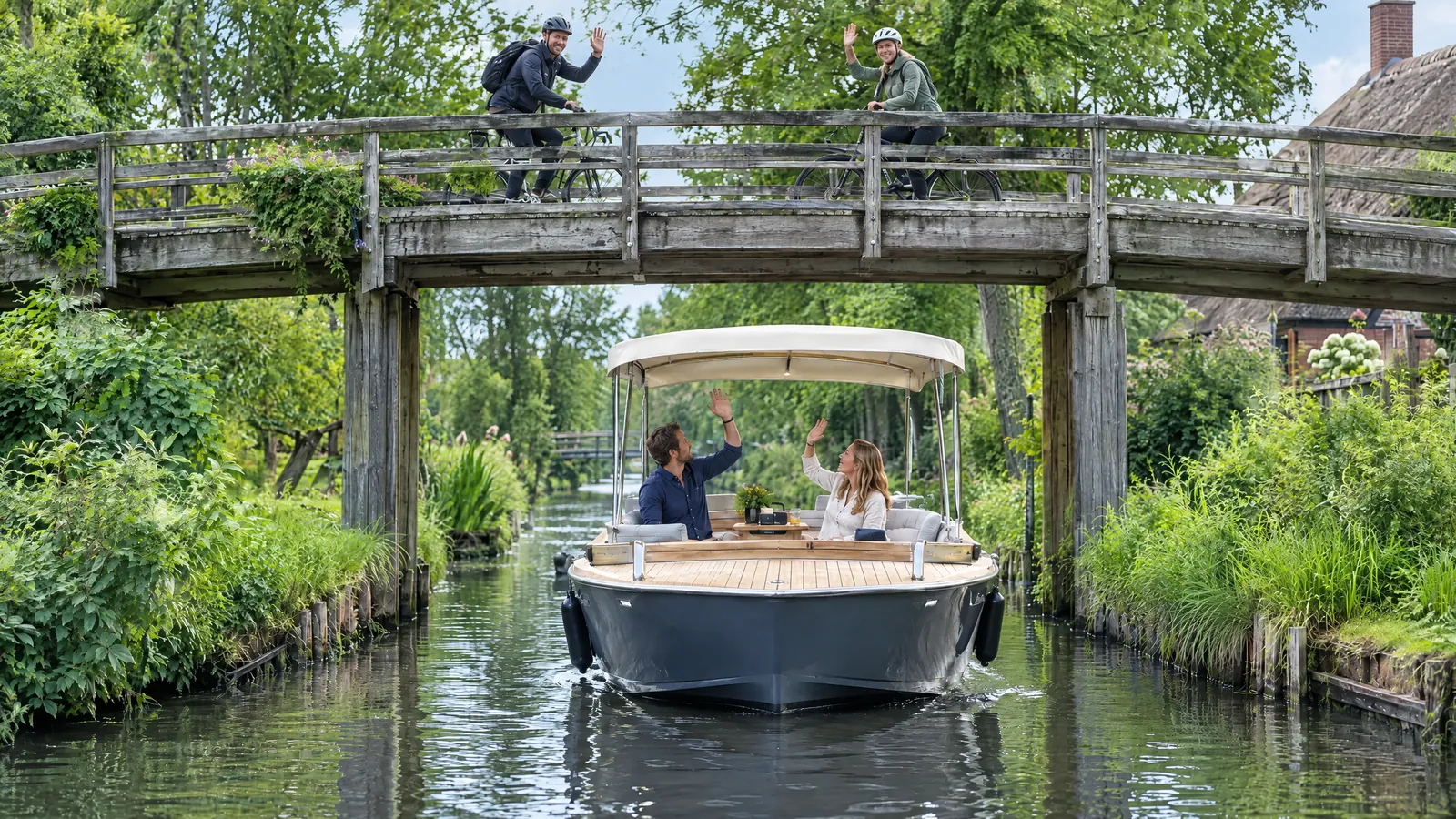 A couple in an electric boat with a sun canopy wave to two cyclists on a wooden bridge